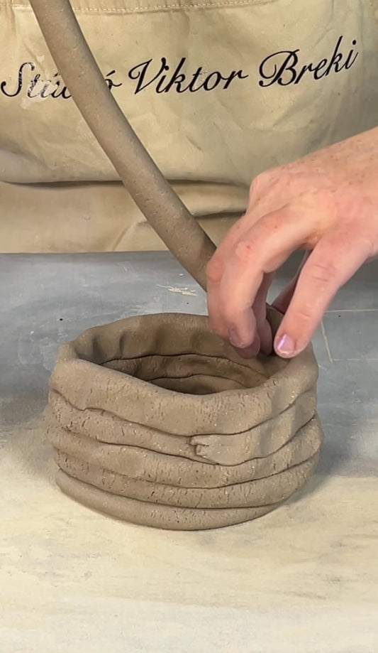 A handmade clay pot with a textured, ribbed exterior, being shaped by a person's hand using a clay lever. The pot is set on a gray work surface with a beige potter's apron in the background.